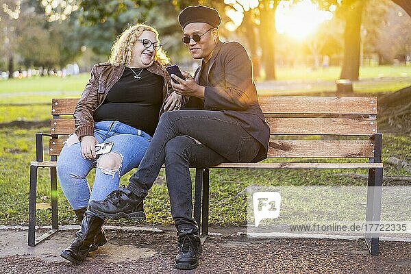 Schwule Freunde sitzen bei Sonnenuntergang auf einer Bank im Park  unterhalten sich und schauen auf ihr Handy