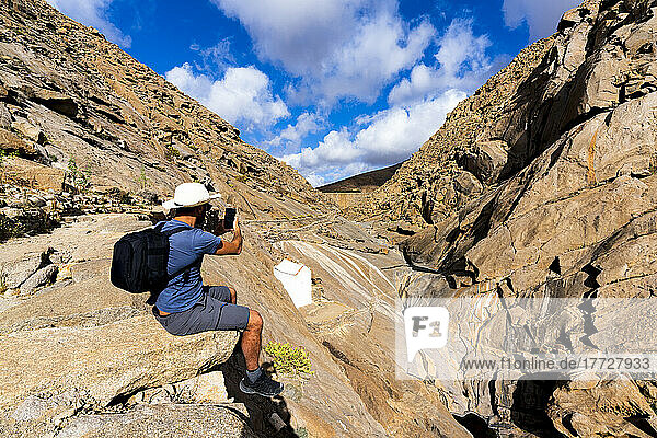 Tourist photographing canyons with smartphone sitting on rocks at Barranco de las Penitas  Fuerteventura  Canary Islands  Spain  Atlantic  Europe