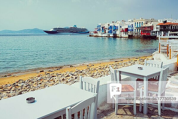 Tourist griechische Szene  Restaurant Café Tisch am Kai Promenade mit Kreuzfahrtschiff und Ägäis und Klein Venedig Häuser im Hintergrund auf schönen Sommertag. Chora  Insel Mykonos  Griechenland  Europa
