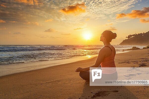 Frau macht Yoga  meditieren und entspannen in Padmasana Lotus Asana Pose mit Kinn Mudra im Freien am tropischen Strand bei Sonnenuntergang mit dramatischen Sonne