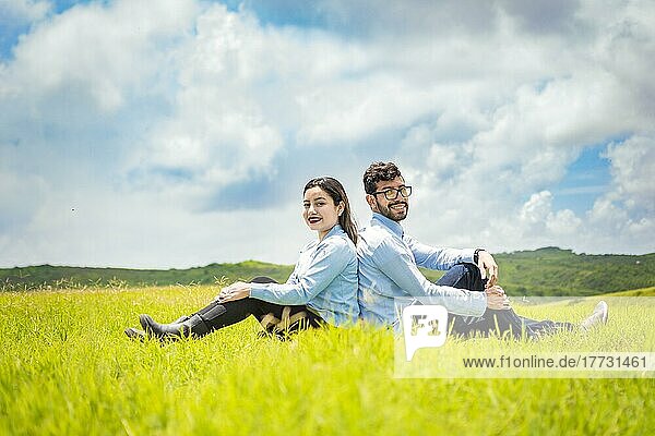 A wedding couple sitting with their backs to each other on the grass  Wedding couple in the field sitting with their backs to each other looking towards the camera