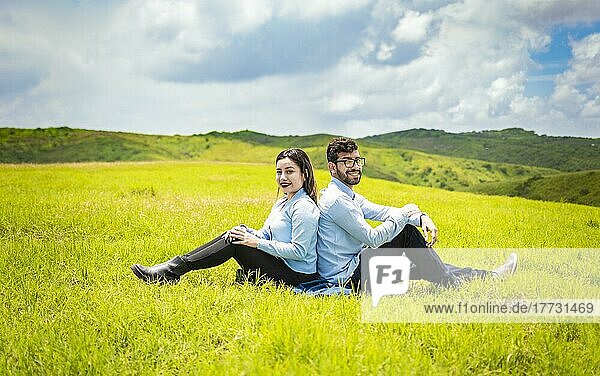 Hochzeitspaar auf dem Feld  Rücken an Rücken sitzend und der Kamera zugewandt  Ein Hochzeitspaar  das Rücken an Rücken auf dem Gras sitzt