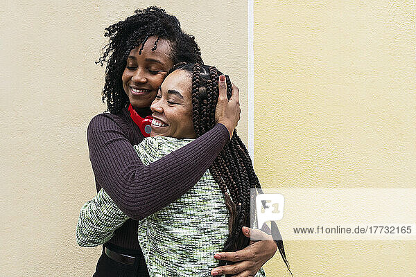 Smiling female friends hugging each other standing in front of wall