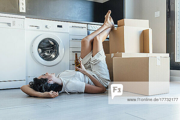 Happy woman holding beer bottle with feet up on cardboard box lying by washing machine in utility room