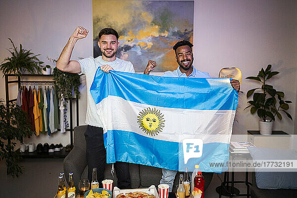 Cheerful young men gesturing fists standing with Argentinian flag at home