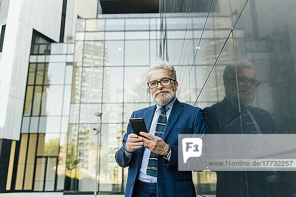 Smiling businessman standing with smart phone by wall outside office building