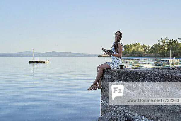 Frau mit Buch sitzt auf Steg am See