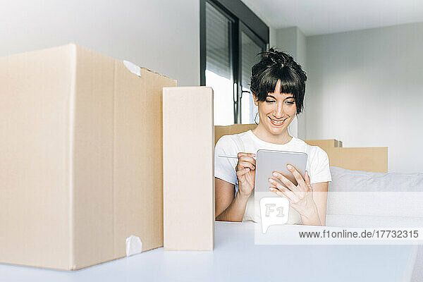 Happy woman using tablet PC sitting with cardboard boxes at table at new home