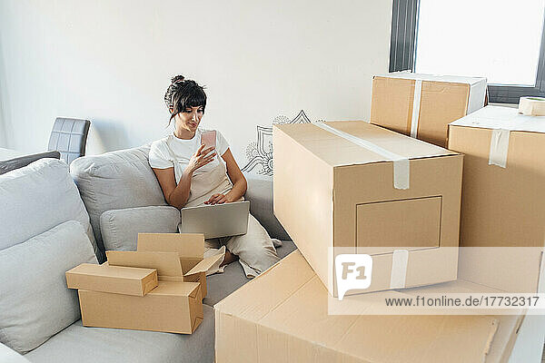Woman using mobile phone sitting with laptop and cardboard boxes in living room