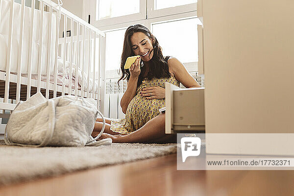 Lächelnde schwangere Frau  die zu Hause am Kinderbett sitzt und Voicemail über ihr Smartphone sendet