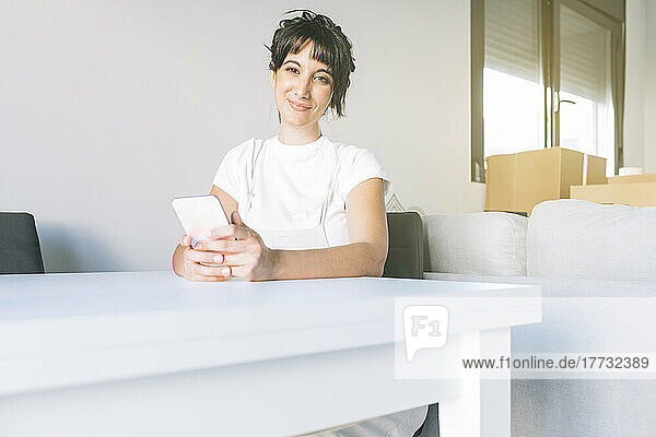 Smiling woman with smart phone sitting at table in living room