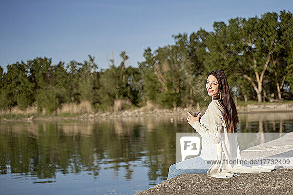 Smiling woman with coffee cup sitting on jetty by lake