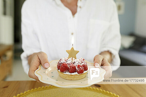 Woman holding plate of raspberry tart with lit candle at home