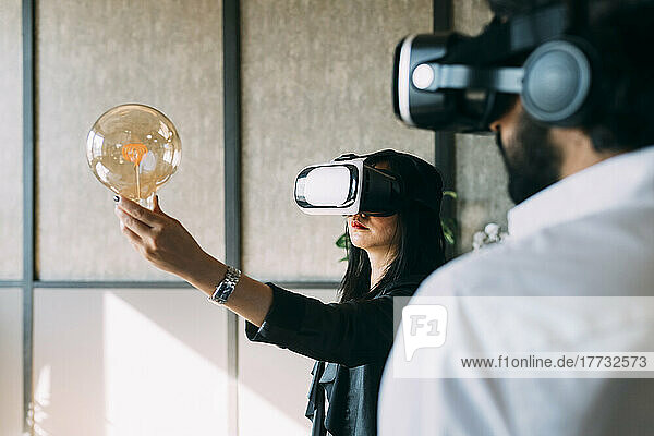 Businesswoman with VR glasses examining light bulb by colleague at office