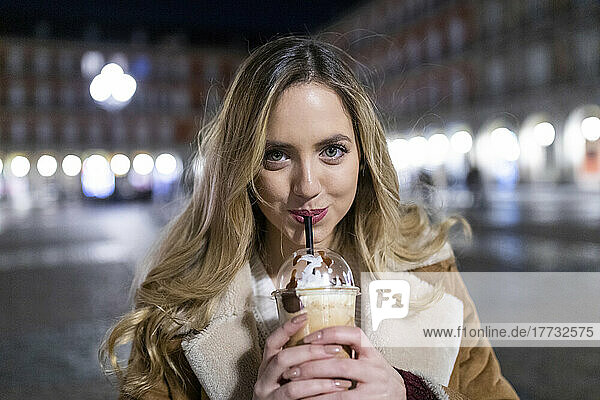 Young woman drinking from disposable cup in city at night