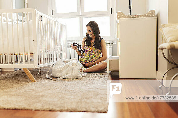 Lächelnde schwangere Frau mit Ultraschalluntersuchungen  die zu Hause am Kinderbett sitzt