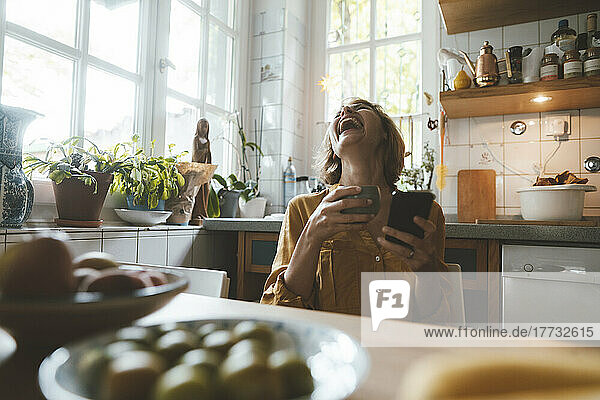 Fröhliche Frau mit Kaffeetasse und Smartphone sitzt am Esstisch