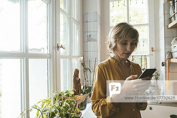 Mature woman using smart phone standing in kitchen