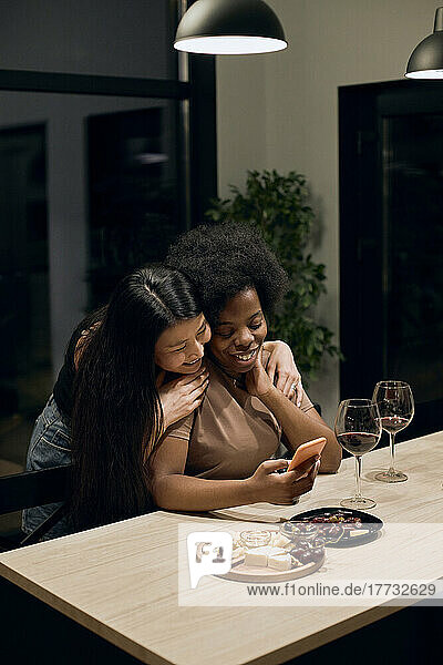 Happy woman embracing girlfriend taking selfie smart phone sitting at table