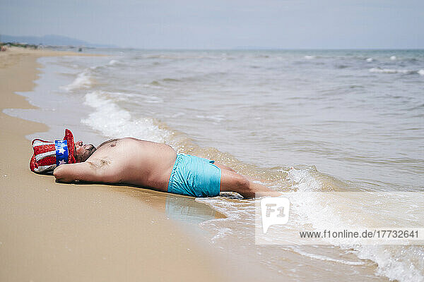 Carefree shirtless man wearing Uncle Sam hat relaxing near seashore at beach