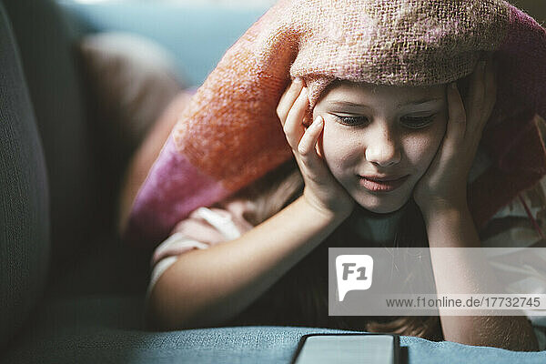 Smiling girl with head in hands using smart phone lying under blanket at home