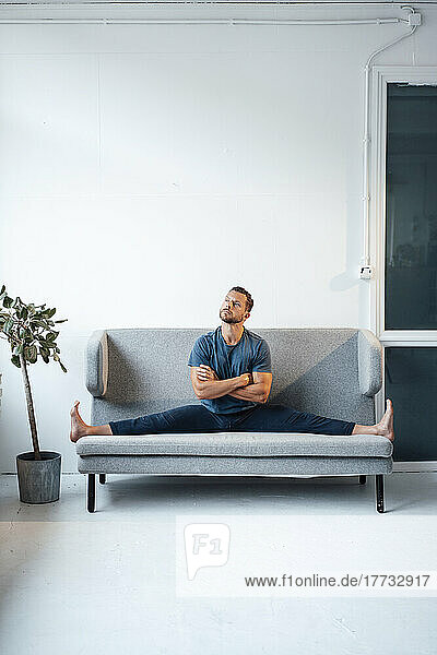 Nachdenklicher Mann streckt die Beine aus und sitzt mit verschränkten Armen zu Hause auf dem Sofa