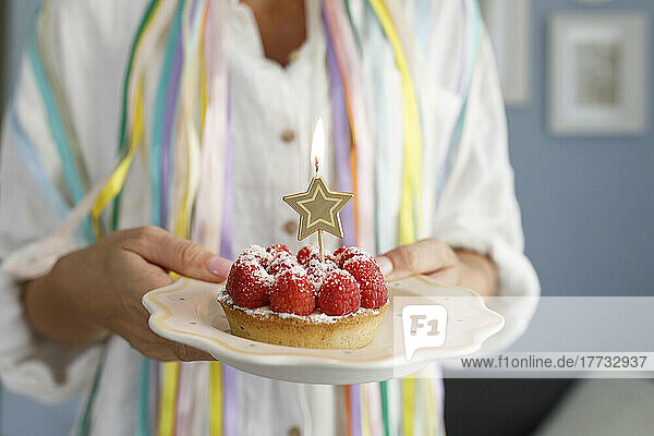 Woman holding plate of raspberry tart with star shaped lit candle at home