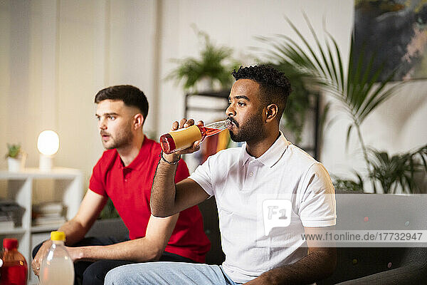 Young man drinking sitting by roommate watching football match at home