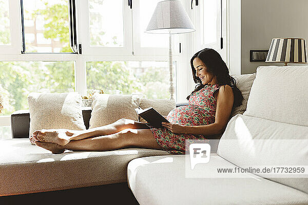 Smiling pregnant woman reading book sitting on sofa in living room at home