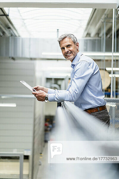 Smiling businessman leaning on railing in factory holding digital tablet