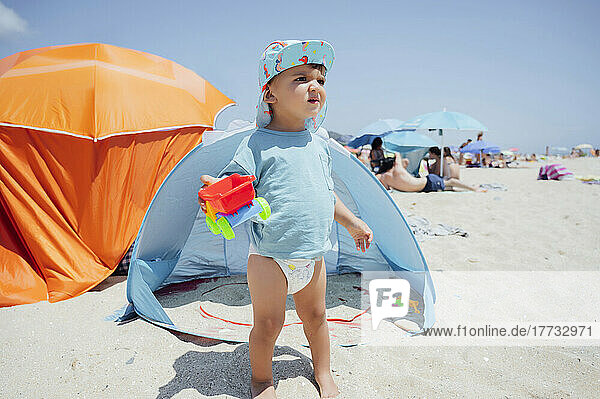 Boy holding toy car standing in front of tent at beach on sunny day