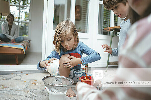 Blond boy poring soil through sieve sitting by friends in front of house