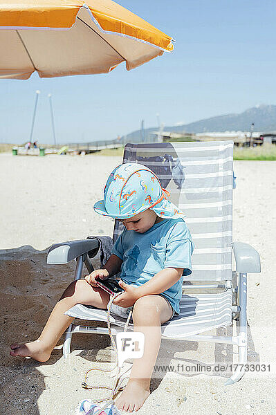 Boy with smart phone sitting on deck chair at beach