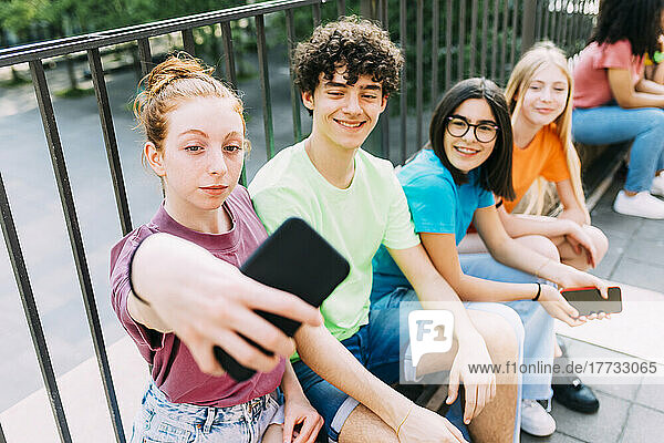 Smiling teenage girl taking selfie with friends sitting by railing