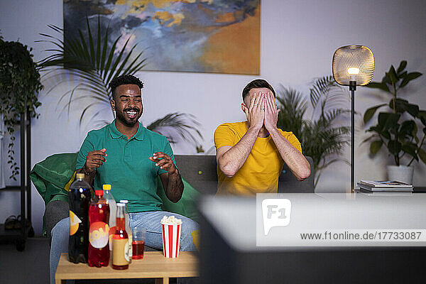 Sad man covering face sitting by happy friend watching football match at home