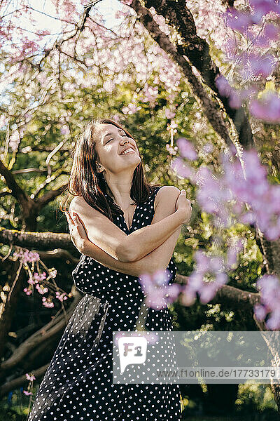 Smiling young woman with eyes closed hugging herself in garden