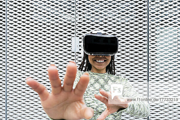 Smiling young woman wearing virtual reality simulator gesturing in front of metal wall