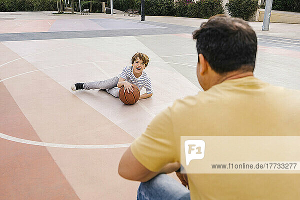 Happy boy with basketball sitting in front of father at sports court