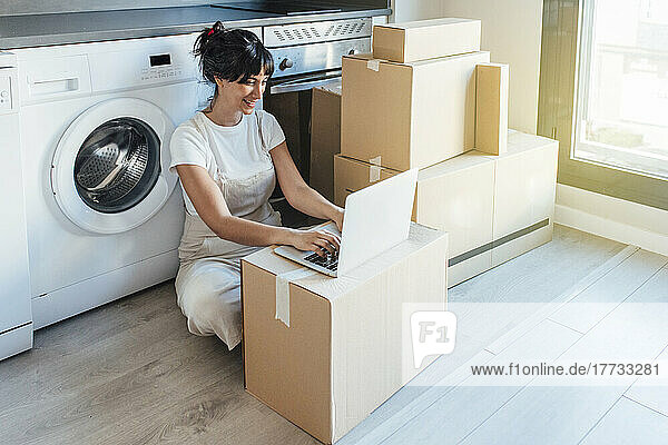 Happy woman using laptop on cardboard box by washing machine in utility room