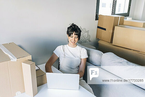 Smiling woman using laptop sitting with cardboard boxes at table in living room