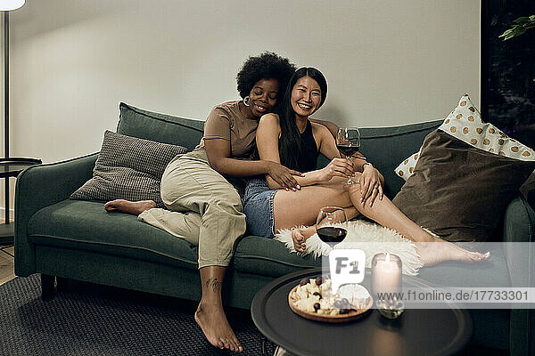 Smiling young woman embracing girlfriend sitting with wineglass on sofa at home
