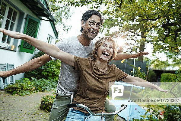 Cheerful mature woman and man with arms outstretched on bicycle in back yard