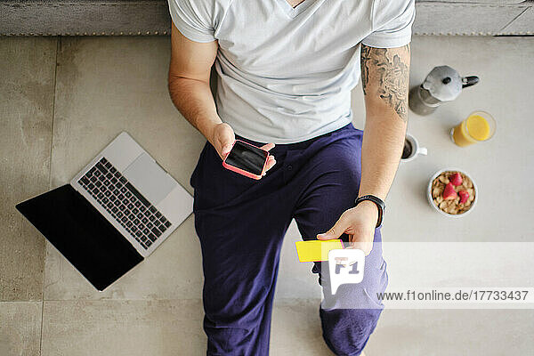 Young man holding credit card doing online shopping through smart phone at home