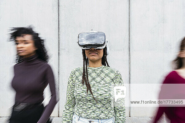 Smiling young woman wearing virtual reality simulator standing in front of wall