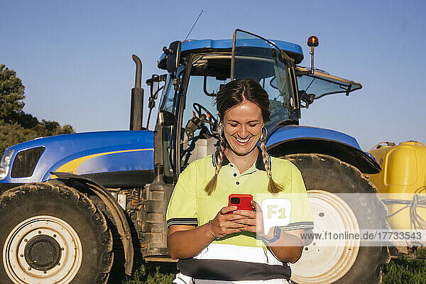 Happy female farmer using smart phone in front of tractor on sunny day