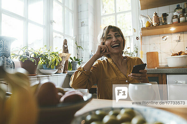 Happy mature woman with smart phone sitting at dining table