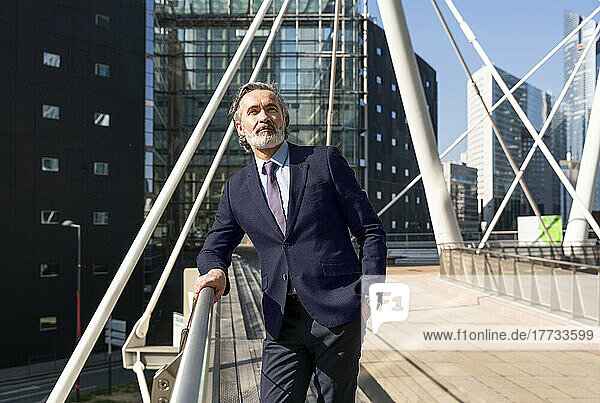 Mature businessman with hand in pocket standing by railing on sunny day