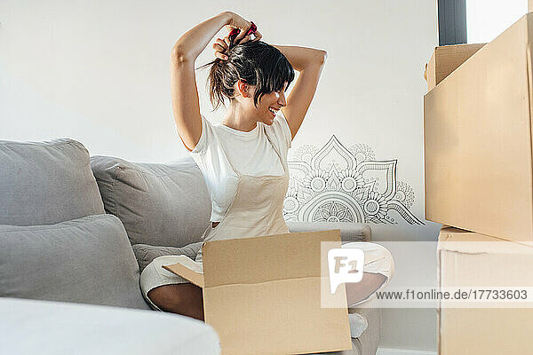 Happy woman tying hair sitting with cardboard box on sofa in living room at home