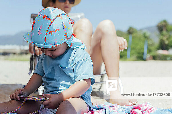 Boy using mobile phone in front of mother relaxing at beach on sunny day