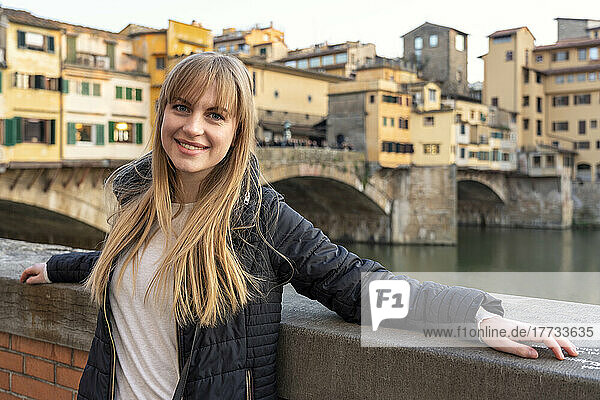 Lächelnde schöne Frau mit Pony steht an der Wand vor der Brücke Ponte Vecchio
